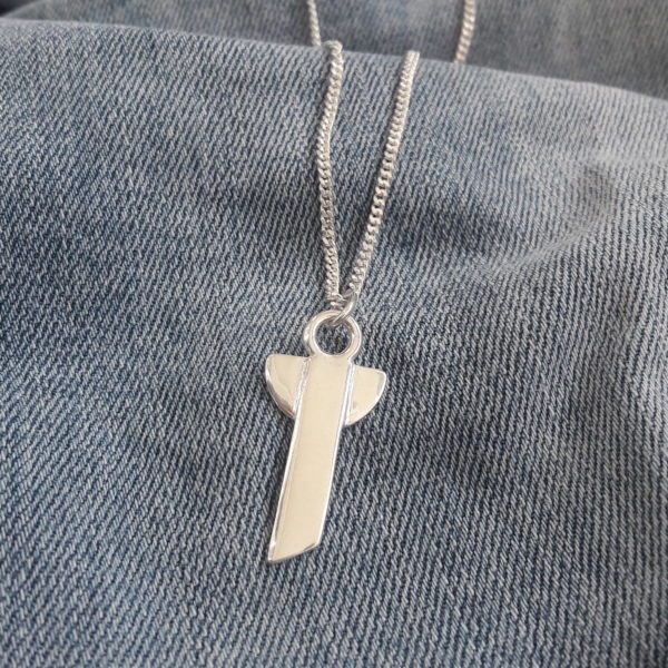 Skruttängel 5 - en stilren silverberlock som kann ses både som ängel och kors med halskedja mot jeansfärgad bakgrund.