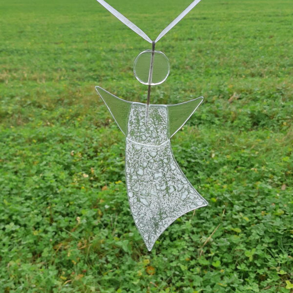 En lätt vitmönstrad ängel av återvunnet fönsterglas - en Gladängel. Hängande i vitt medföljande band mot grön bakgrund. Ca 20 cm.